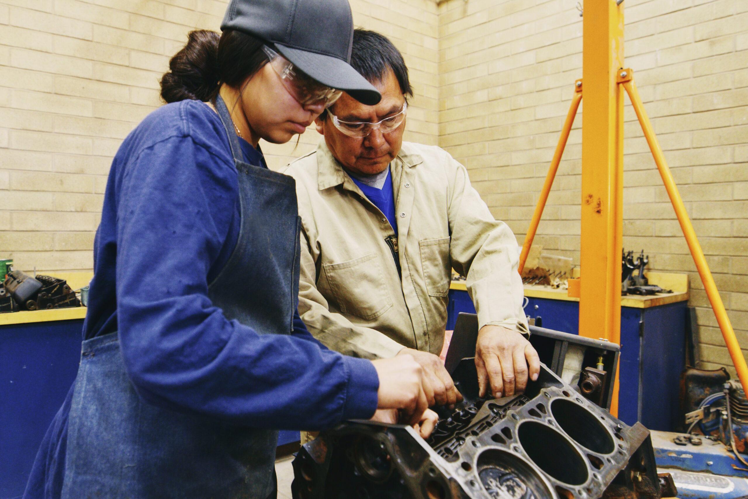 A young Indigenous Navajo woman, working with a teacher in an automotive shop class at a high school.
